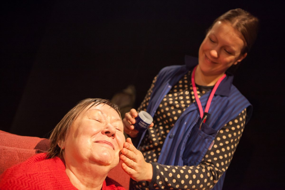 Teatteriesitys, jossa naisnäyttelijä koskettaa toista naisnäyttelijää poskesta, näyttelijät hymyilevät.