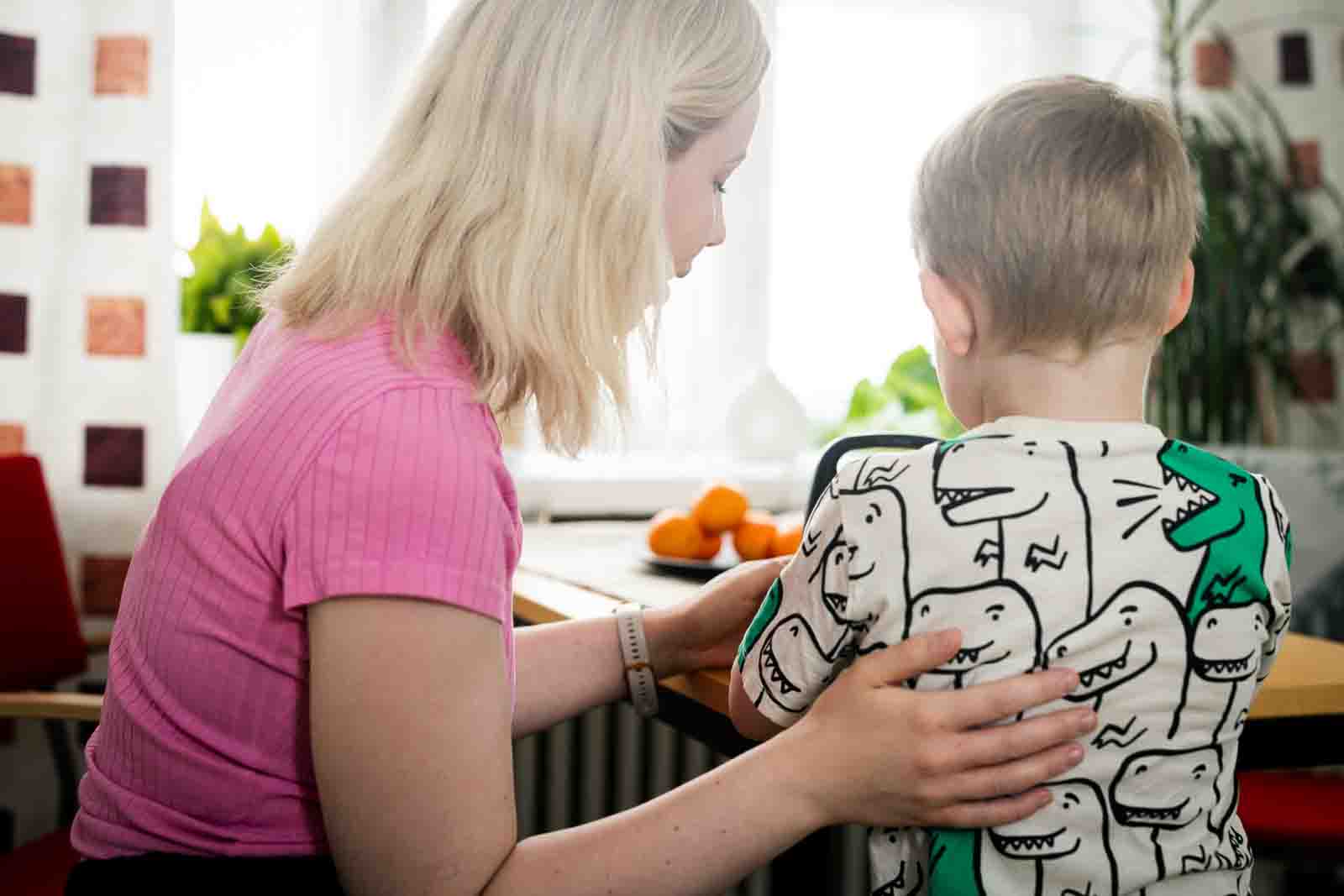 Lapsi ja työntekijä ovat pöydän ääressä. Työntekijä ohjaa lasta harjoituksessa. Kuva on otettu takaapäin.