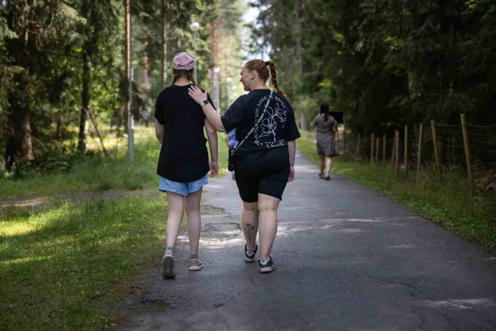 Kaksi naista kävelee kuvaajasta poispäin kujaa pitkin. Kujaa reunustavat puut.