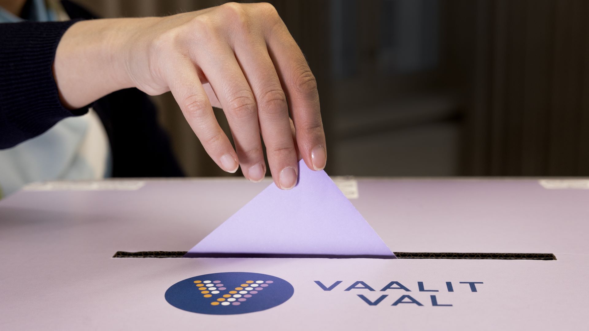 Henkilö tiputtaa violetin äänestyslapun vaaliuurnaan. Vaaliuurnassa lukee Vaalit, Val. Kuvassa henkilöstöstä näkyy käsi.