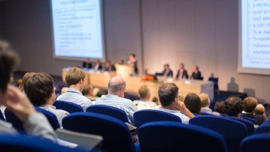 Ihmiset istuvat auditoriossa. Edessä, keskellä on puhujan korokkeella yksi henkilö ja ympärillä istuu seitsemän henkilöä.