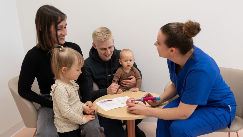 Siniasuinen nainen näyttää näyttöhampaiden avulla, miten hampaita pestään. Etualalla nainen ja pikkutyttö sekä taka-alalla mies sylissään vauva seuraavat opastusta.