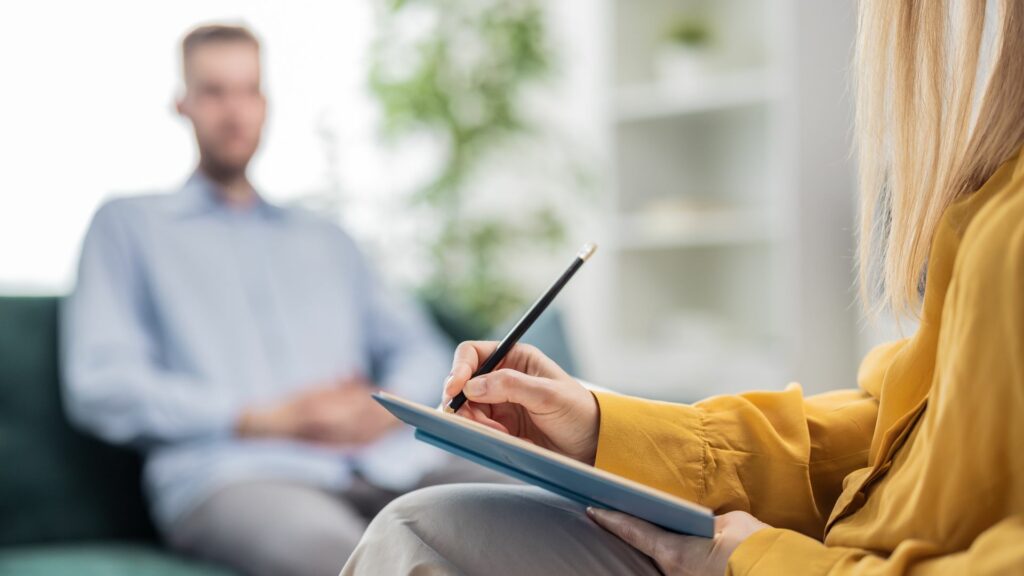Nainen kirjoittaa etualalla muistikirjaan. Mies istuu taustalla sohvalla.